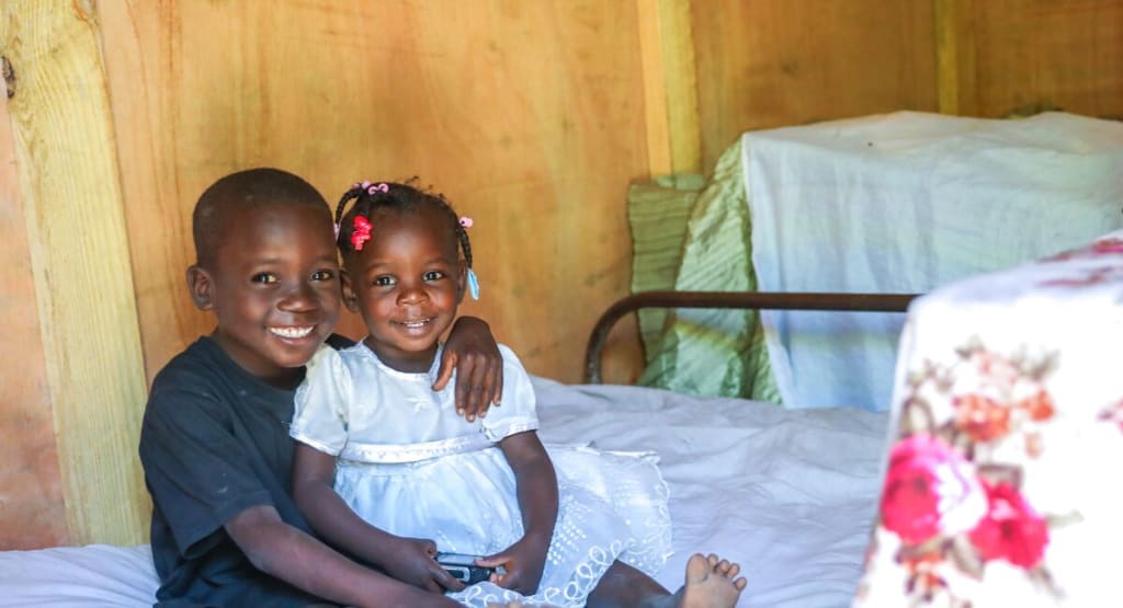 A little girl in a white dress and a little boy in a black t-shirt sit on a bed and smile